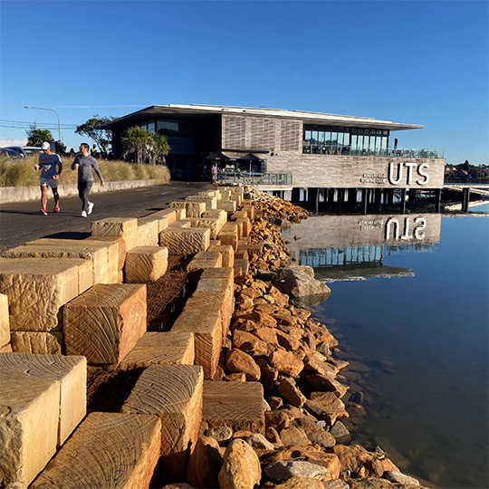 A walking path located on the shore of a bay with a rowing club in the background. The path is raised above a seawall made from both natural and rectangular-shaped sandstone blocks.